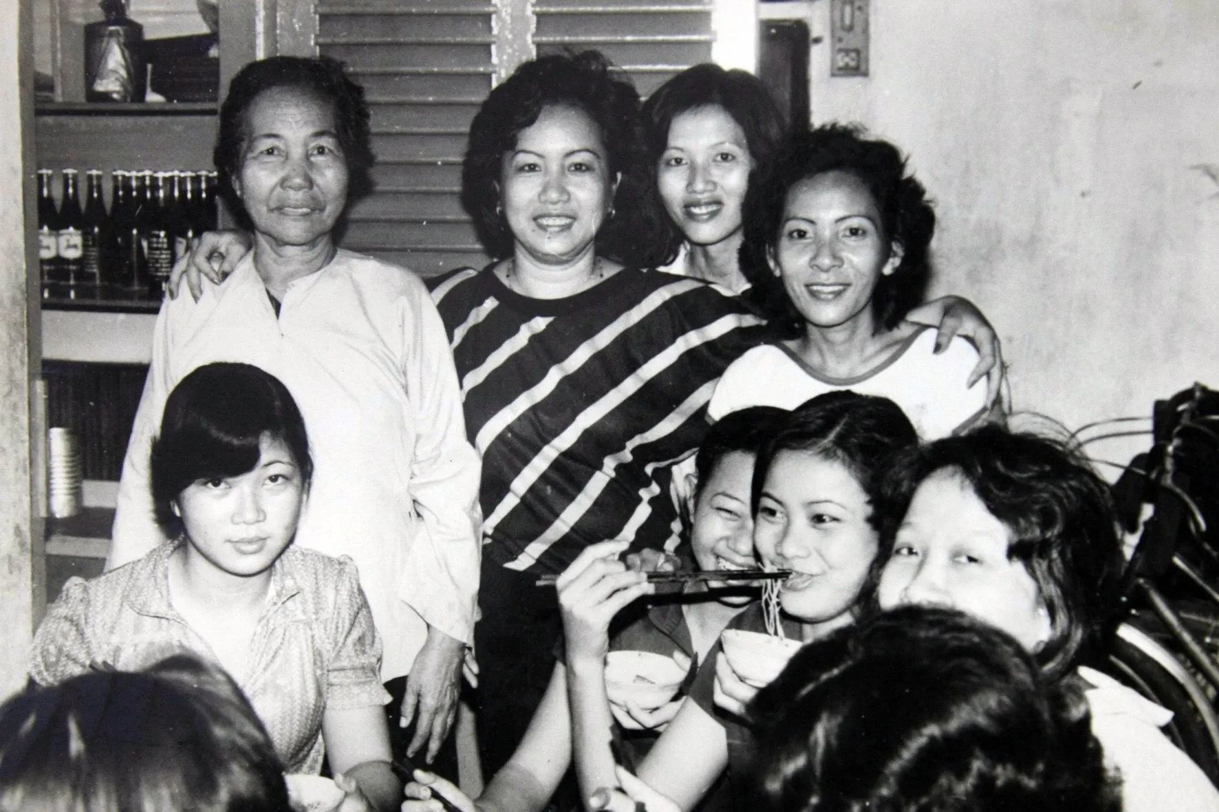 Black-and-white snapshot of smiling women gathered around a table, one eating noodles with chopsticks