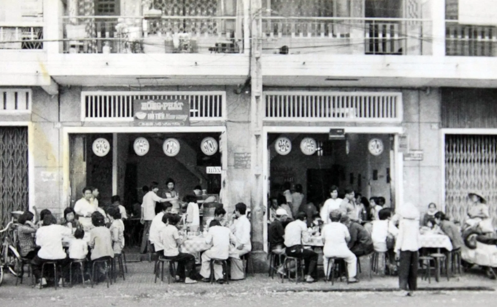 Black-and-white photo of diners on low stools at sidewalk tables outside two Hồng Phát shopfronts