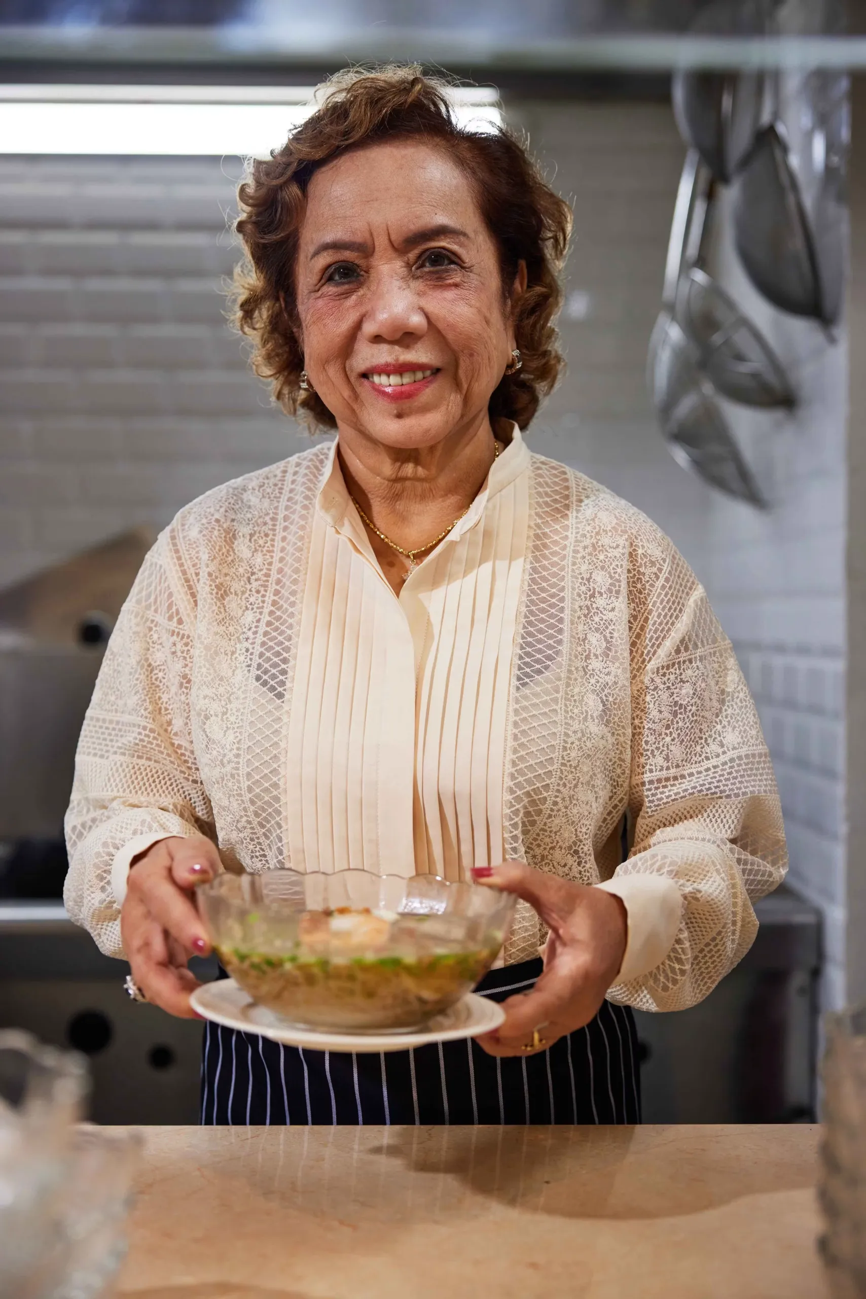 Chef Đặng Thị Nguyệt smiling in a restaurant kitchen, holding a glass bowl of hủ tiếu Nam Vang