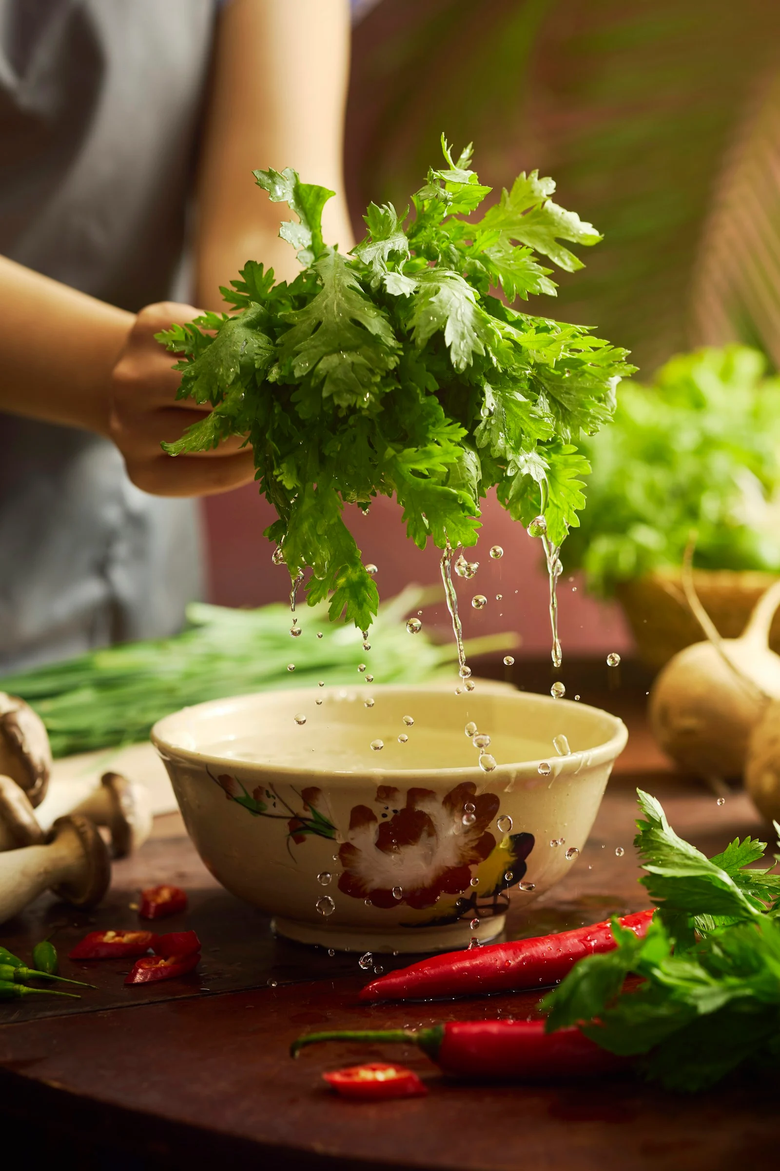 Fresh chrysanthemum greens rinsed over a hand-painted bowl, with red chilies on a wooden table