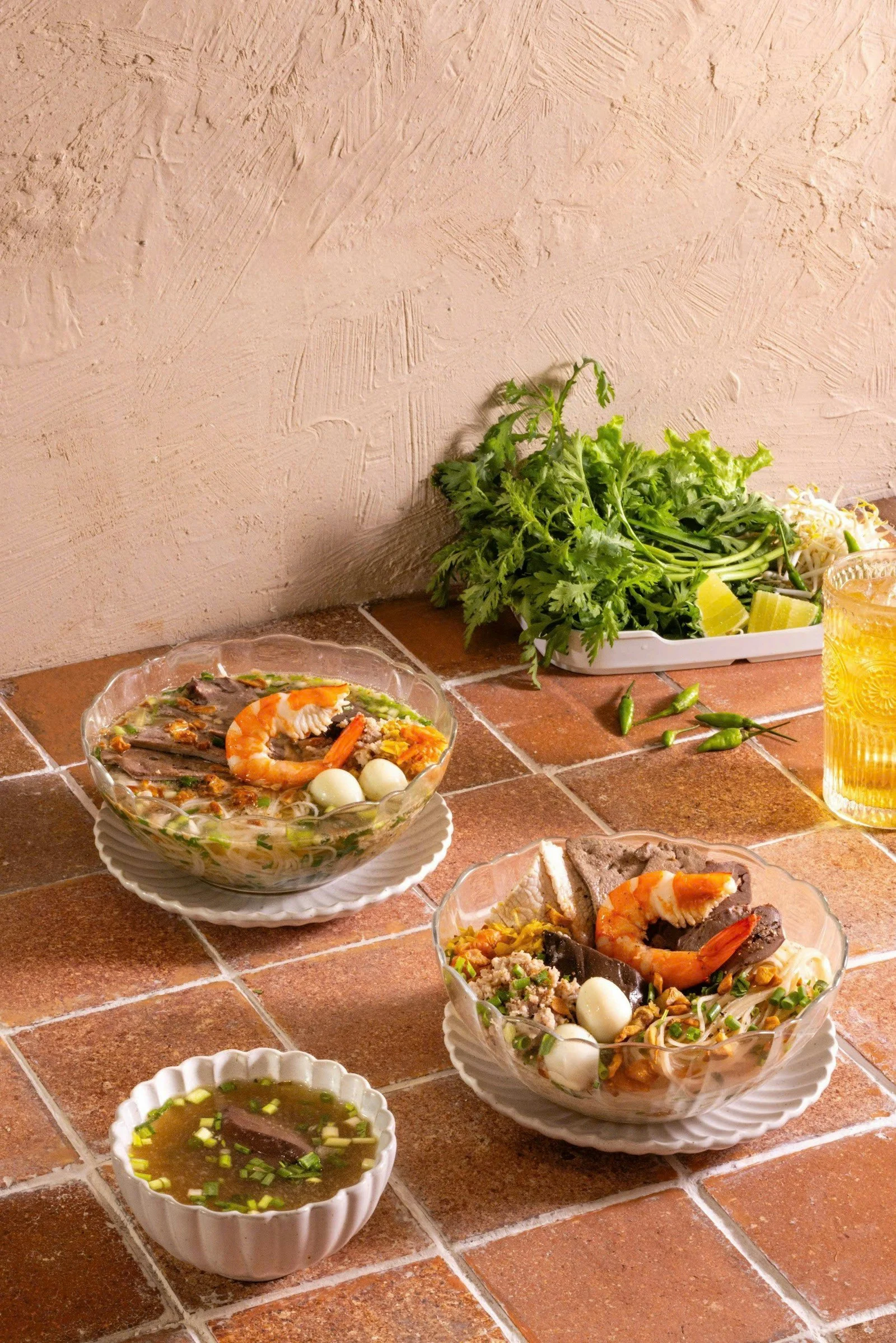 Two bowls of hủ tiếu Nam Vang topped with shrimp, pork, and quail eggs, served with fresh herbs